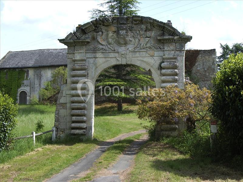 Portail en pierre sculptée donnant accès à une allée bordée de végétation, avec un bâtiment ancien en arrière-plan.