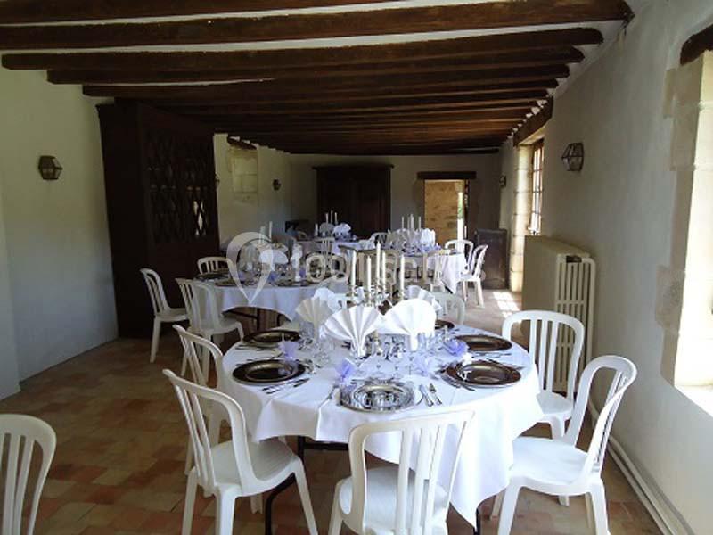 Salle de réception avec tables rondes dressées, nappes blanches, chaises en plastique et poutres apparentes au plafond.