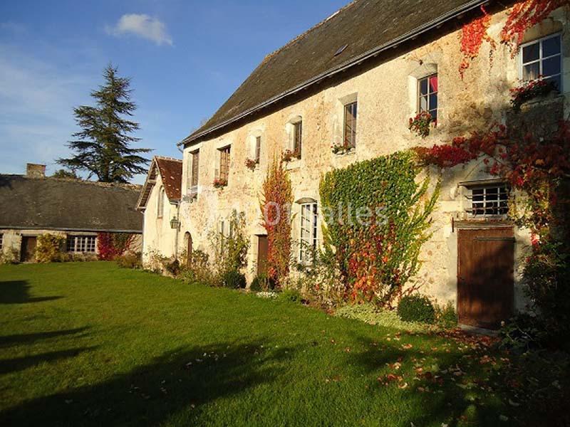 Façade d'une maison ancienne en pierre avec des volets et des plantes grimpantes, entourée d'une pelouse verte.