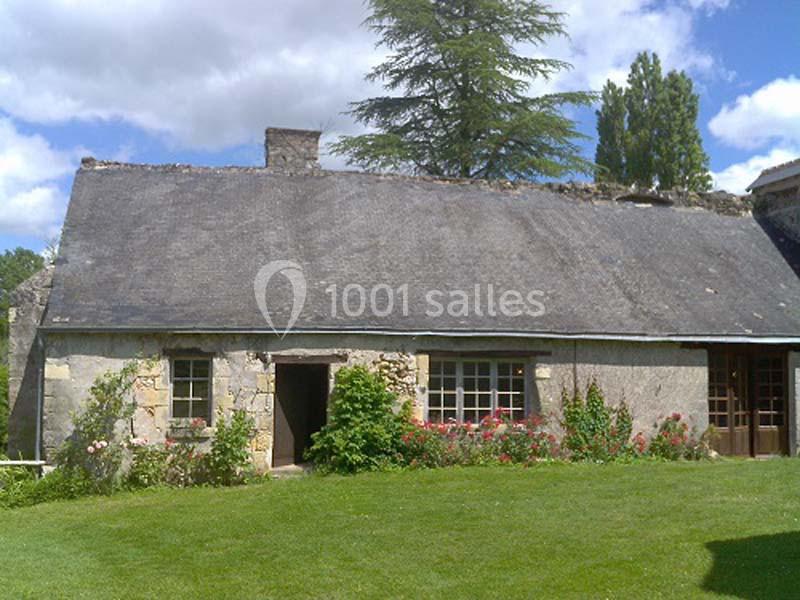 Maison en pierre avec toit en ardoise, entourée de pelouse et de fleurs, sous un ciel partiellement nuageux.