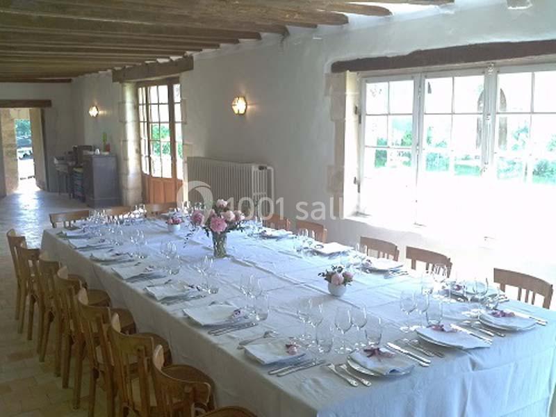 Grande table dressée avec nappes blanches, verres et couverts, dans une salle lumineuse avec poutres apparentes et fenêtres.