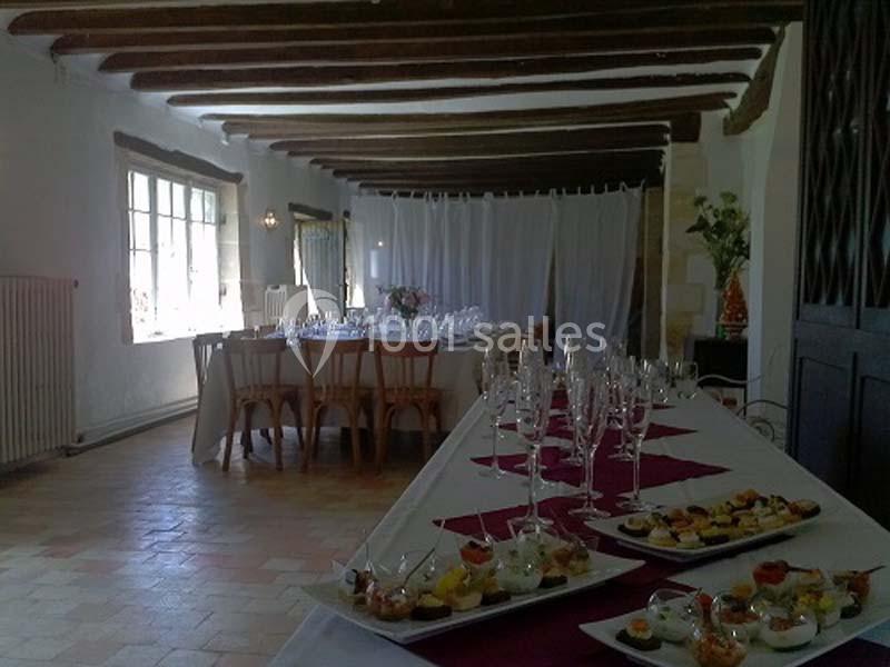 Salle lumineuse avec poutres apparentes, tables dressées avec verres et assiettes de bouchées apéritives.