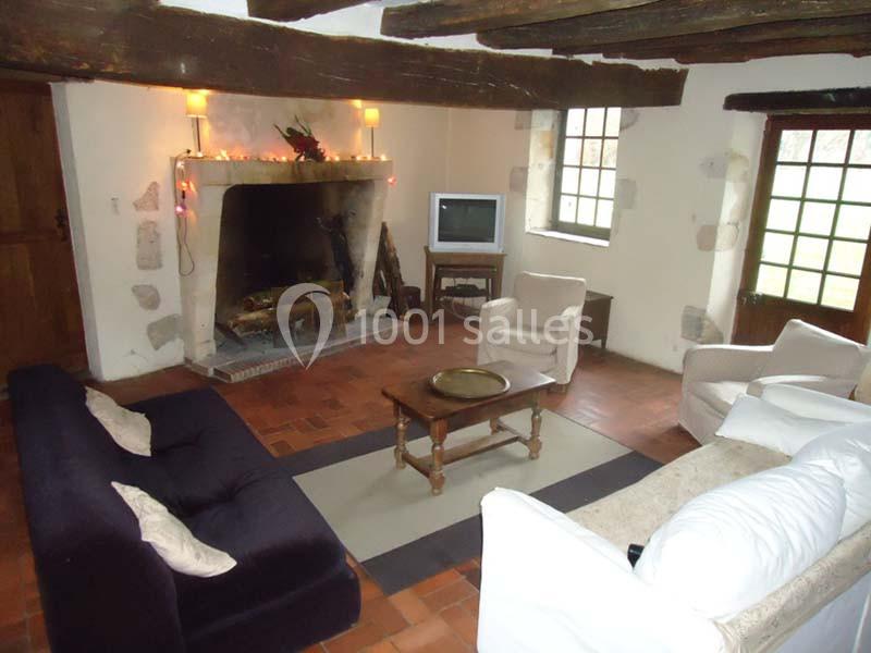 Salon rustique avec poutres apparentes, cheminée en pierre, canapé, fauteuils et table basse sur un sol en terre cuite.