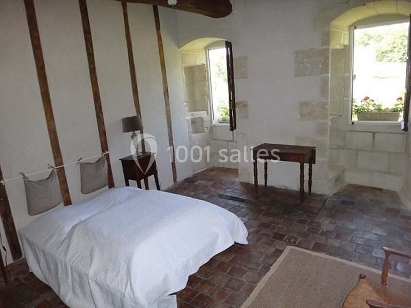 Chambre lumineuse avec murs en pierre, poutres apparentes, sol en carreaux anciens, lit simple et mobilier en bois.