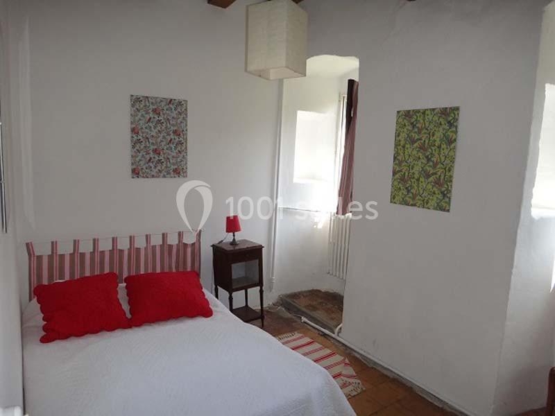 Chambre lumineuse avec lit double, coussins rouges, table de chevet, lampe et décorations murales colorées.