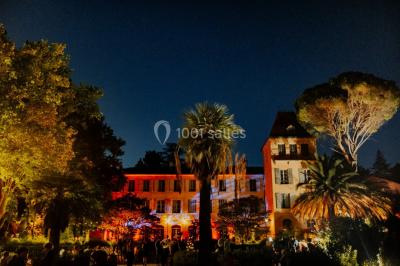 Allée bordée de palmiers éclairés, menant à une fontaine illuminée dans un jardin au crépuscule.