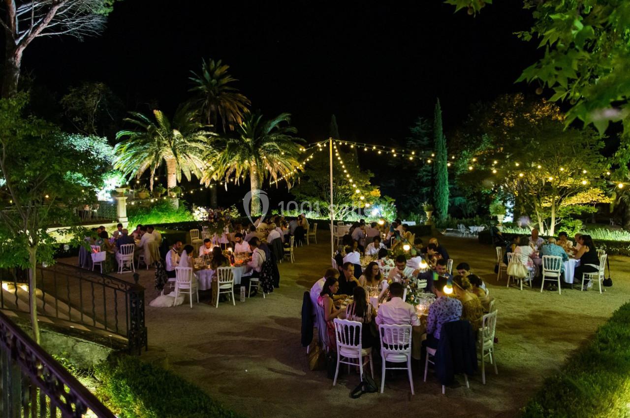 Des convives dînent en plein air dans un jardin éclairé par des guirlandes lumineuses, entouré de palmiers et de verdure.