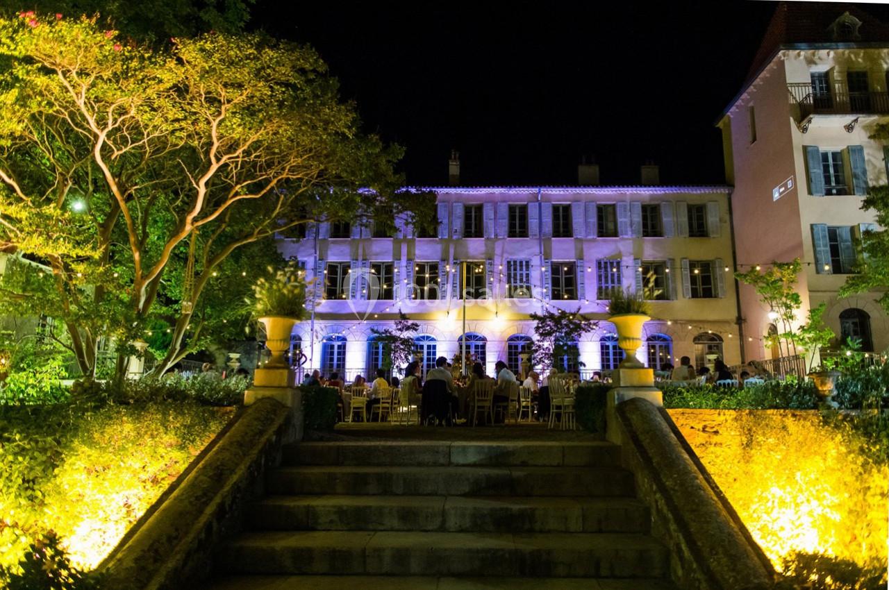 Façade d'un bâtiment éclairé la nuit, entouré de jardins avec des invités assis à des tables en extérieur.