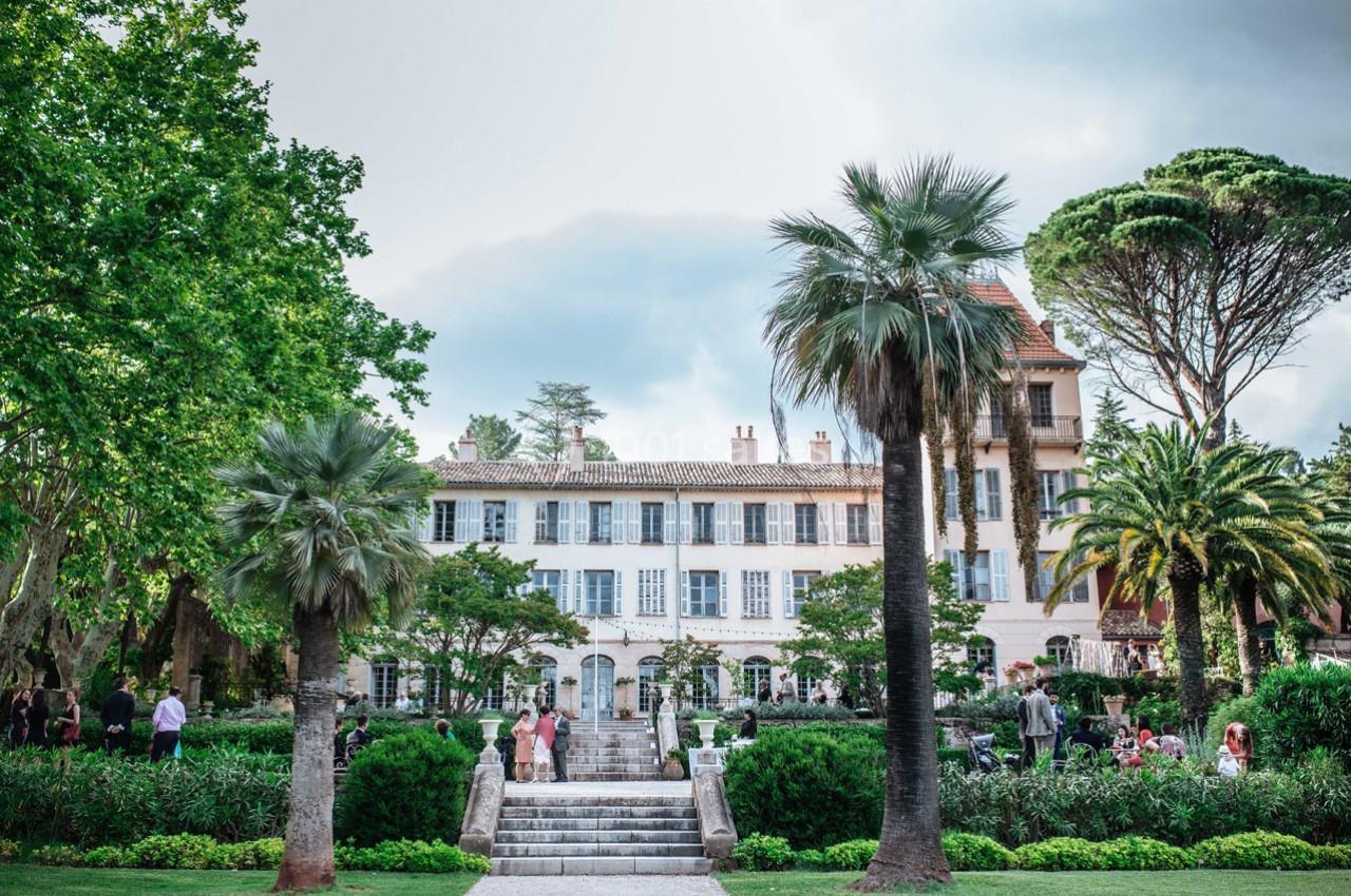 Façade d'une grande maison entourée de jardins verdoyants avec des palmiers et des personnes se promenant.