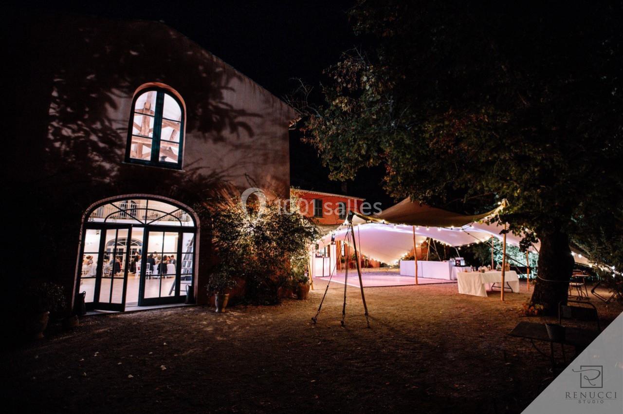 Bâtiment en pierre éclairé de nuit avec une tente lumineuse installée dans un jardin adjacent.