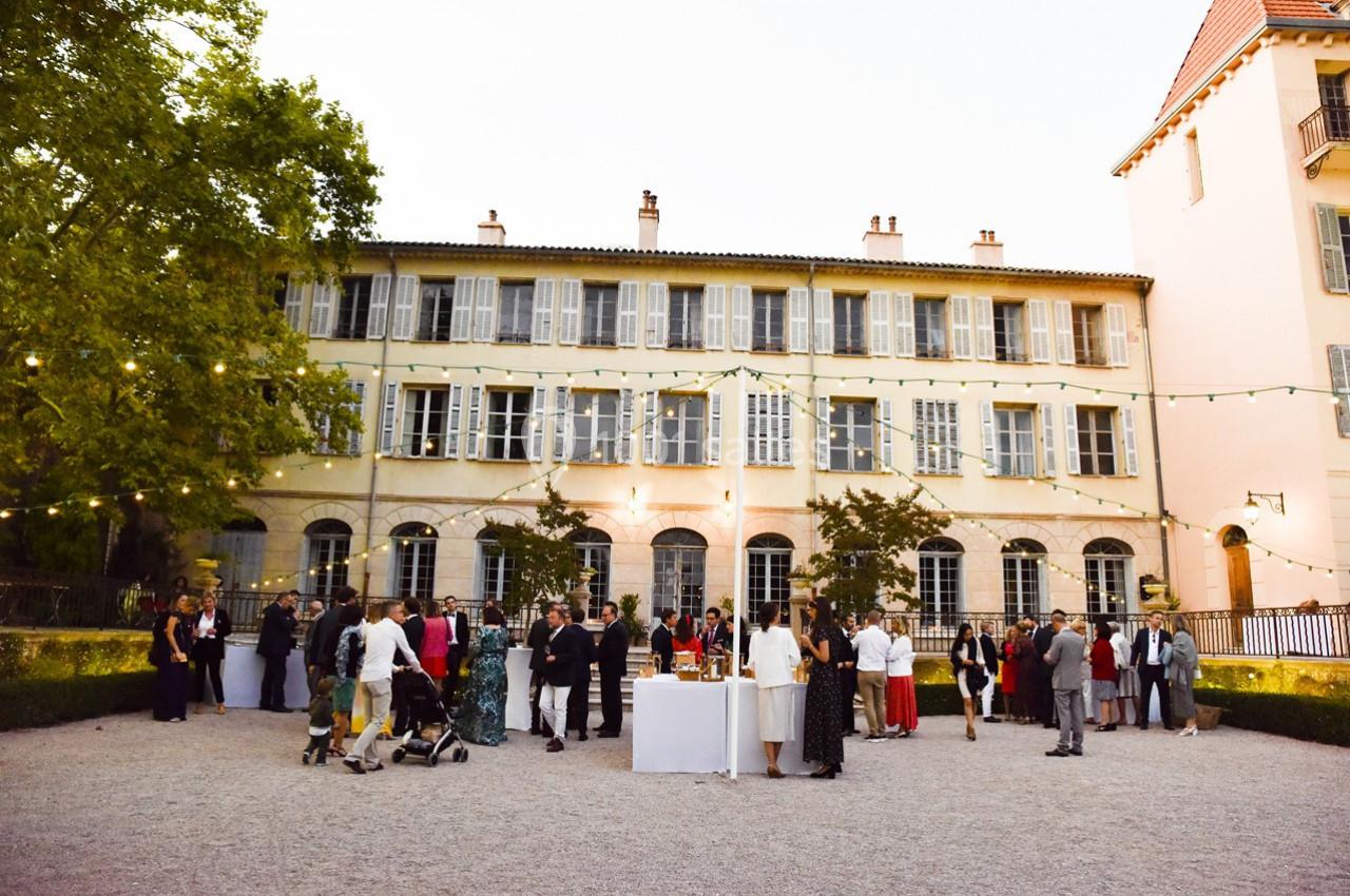 Groupe de personnes rassemblées dans une cour extérieure, devant un grand bâtiment ancien, sous des guirlandes lumineuses.