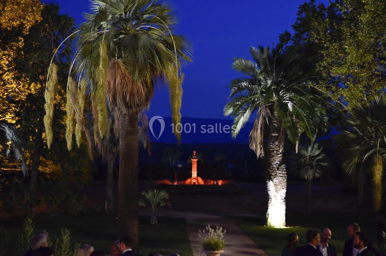 Allée bordée de palmiers éclairés, menant à une fontaine illuminée dans un jardin au crépuscule.