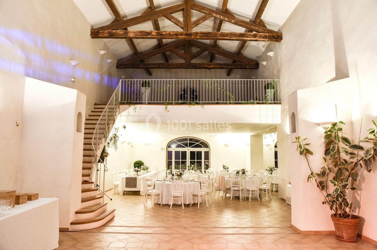 Salle de réception lumineuse avec tables dressées, escalier en colimaçon et poutres apparentes au plafond.