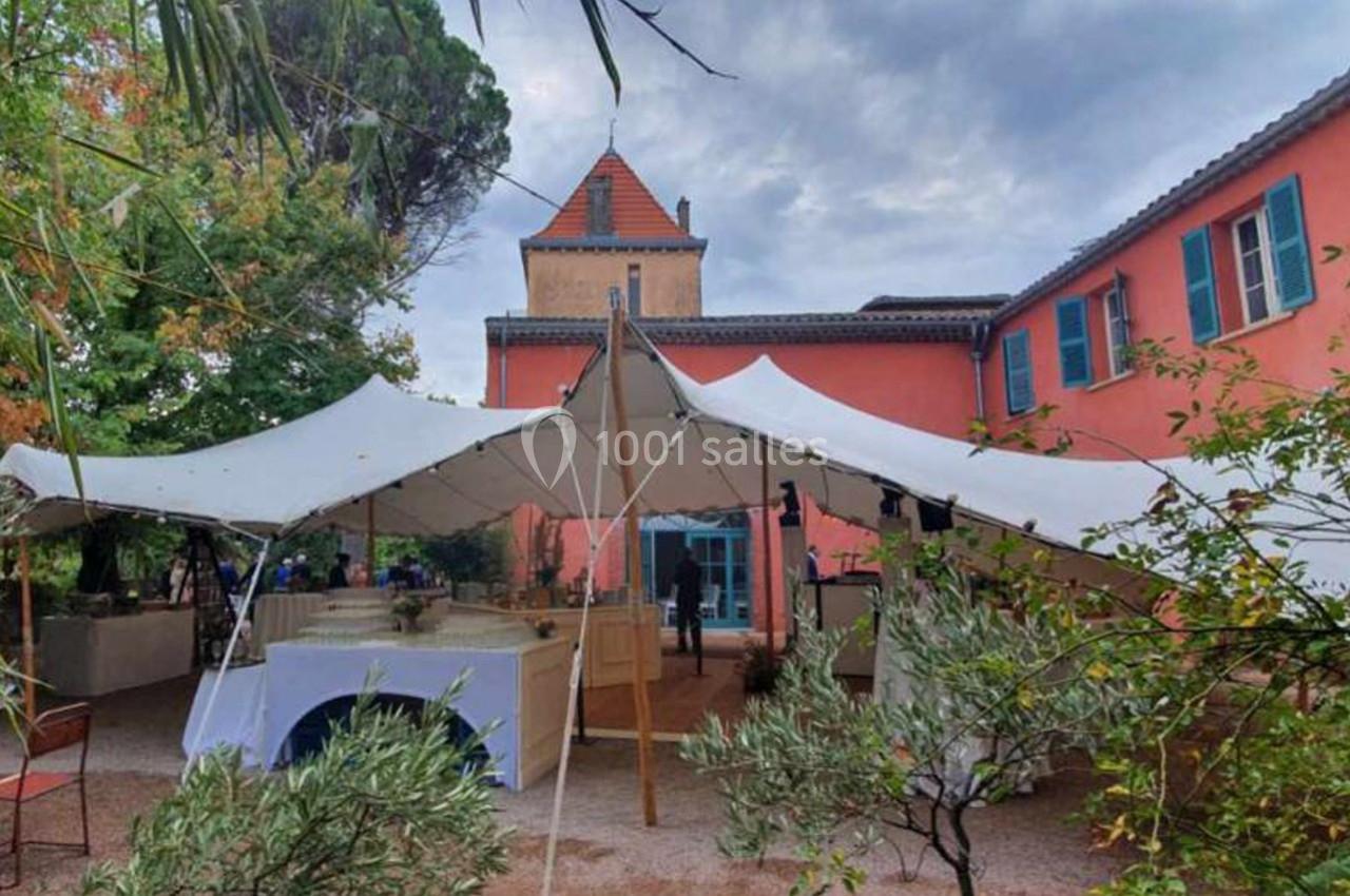 Tente blanche installée dans une cour extérieure devant un bâtiment rose à volets bleus, entouré de végétation.