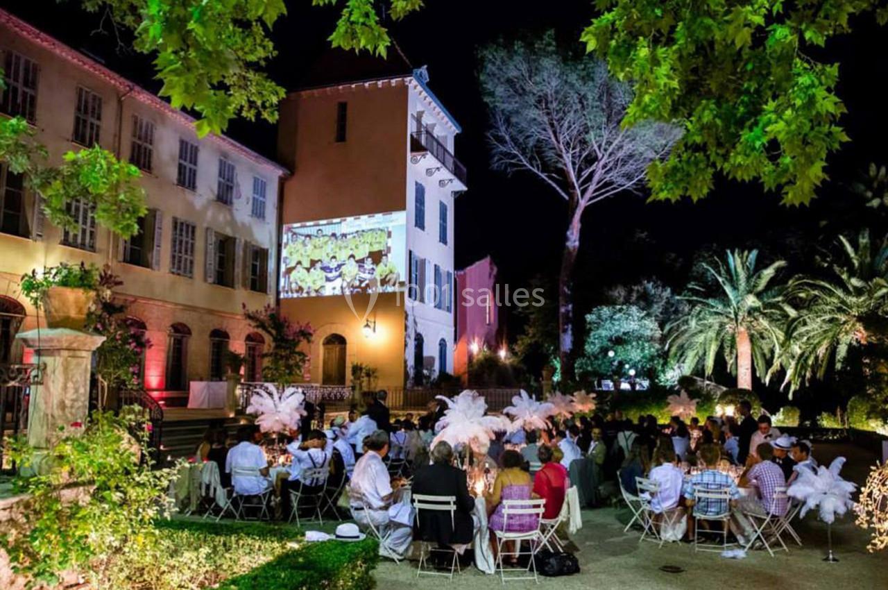 Personnes dînant en plein air dans un jardin éclairé, avec un bâtiment ancien et une projection murale en arrière-plan.