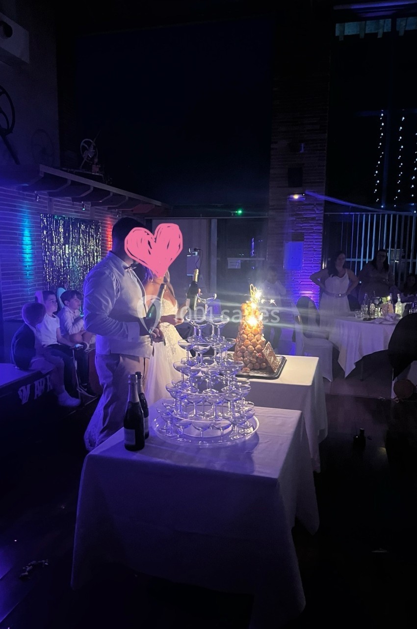 Un homme en tenue blanche près d'une fontaine de verres et d'une pièce montée éclairée dans une salle sombre.