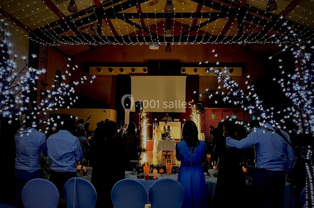 Salle de réception décorée avec des guirlandes lumineuses, des invités debout face à une scène éclairée.