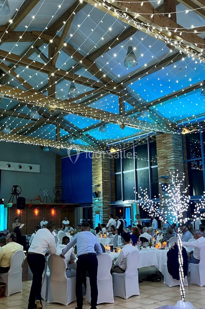 Salle de réception décorée avec des guirlandes lumineuses, des tables dressées et des invités en tenue de soirée.