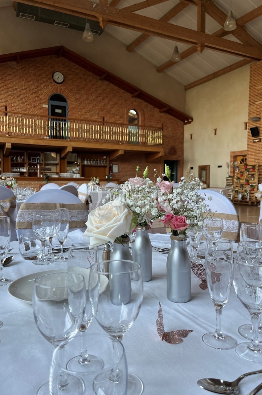 Salle de réception décorée pour un événement, avec tables dressées, fleurs et chaises ornées de rubans.