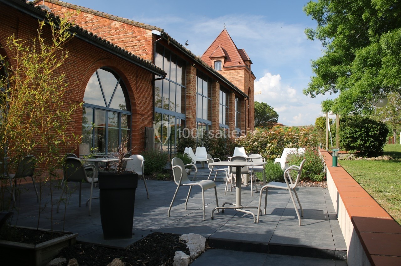 Terrasse extérieure avec tables et chaises, bordée de plantes, devant un bâtiment en briques sous un ciel dégagé.
