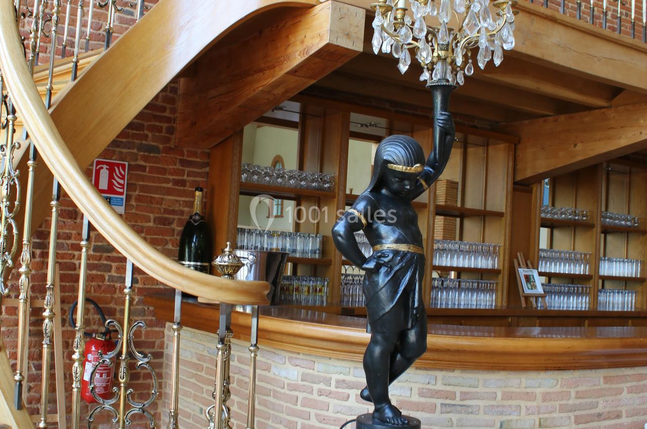 Escalier en bois courbé avec balustrade dorée, statue décorative sur socle et lustre suspendu devant un mur en briques.