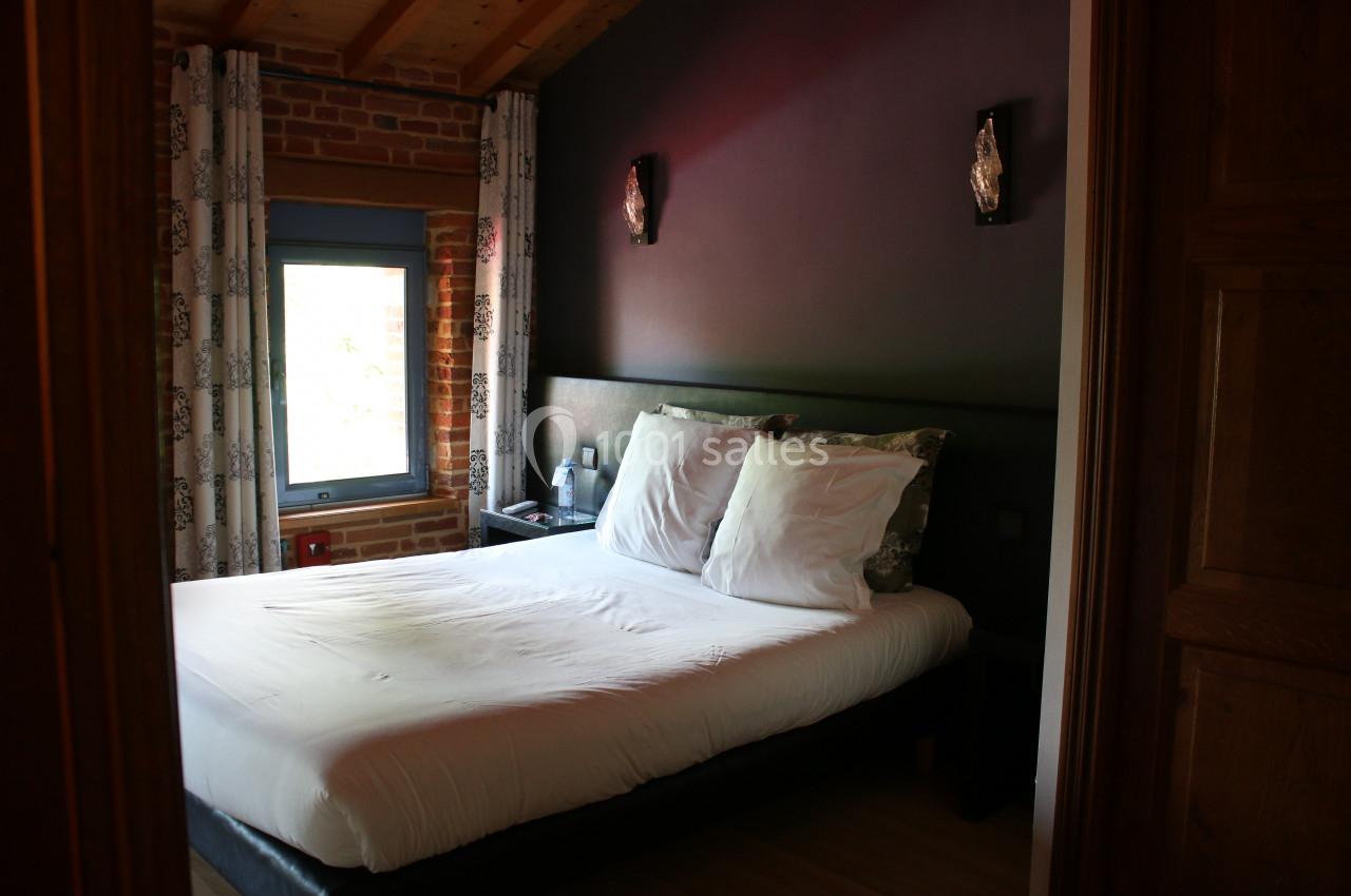 Chambre avec un lit double, draps blancs, murs sombres, fenêtre avec rideaux et éclairage mural.