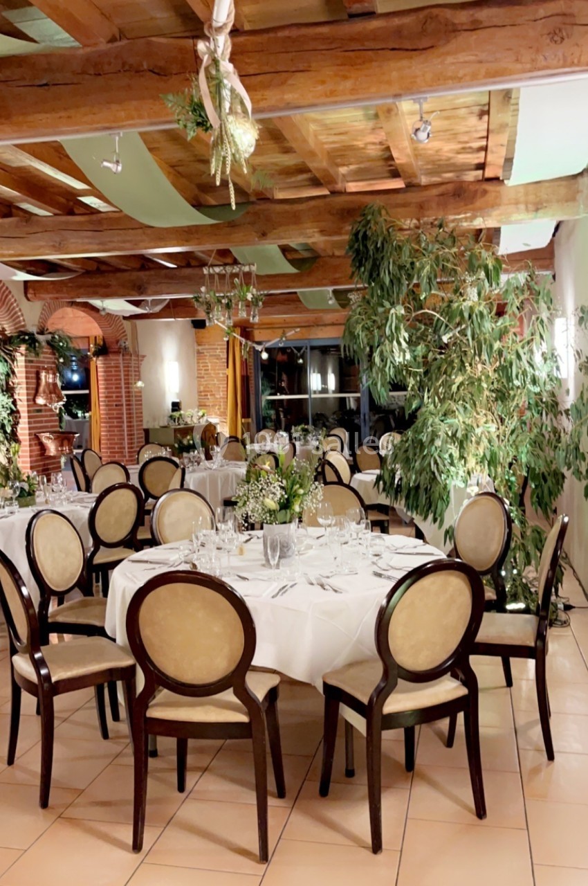 Salle de réception élégante avec tables rondes dressées, chaises en bois, plantes et plafond en poutres apparentes.