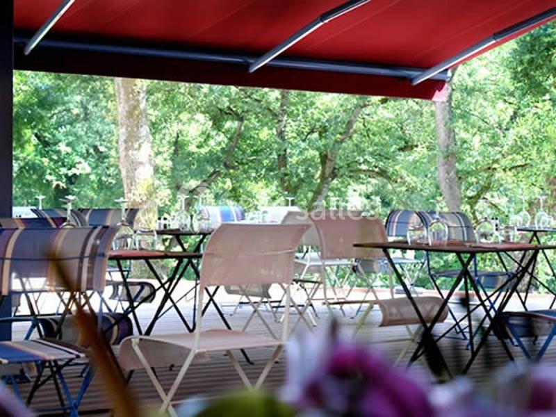Terrasse extérieure avec tables et chaises sous un auvent rouge, entourée d'arbres et de verdure.