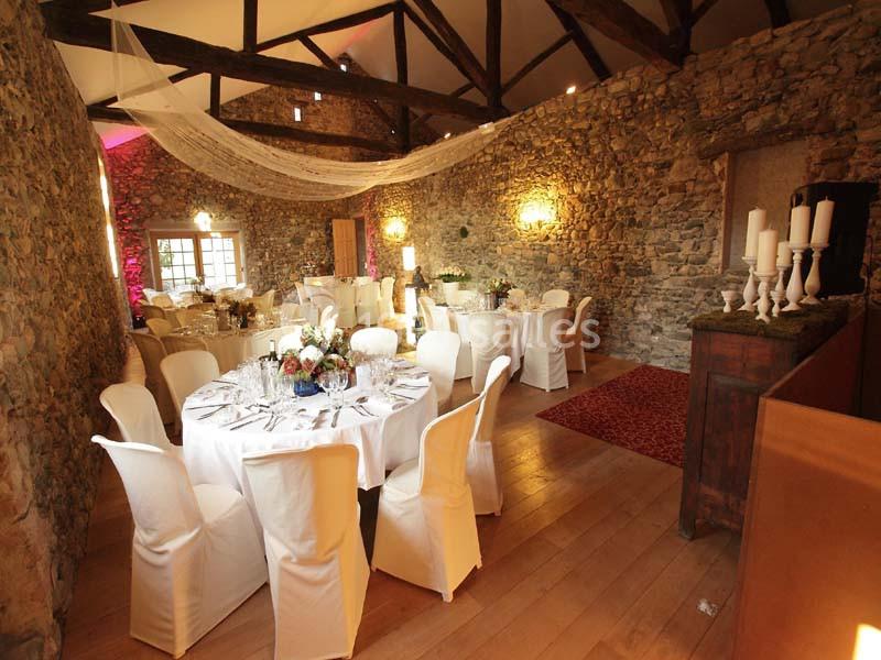 Salle en pierre avec poutres apparentes, tables rondes dressées pour un événement, décorations élégantes et lumière tamisée.