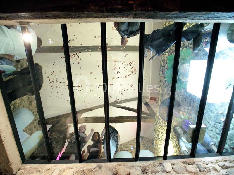 Vue depuis un sol en verre montrant des personnes debout au-dessus, dans un intérieur avec poutres apparentes.