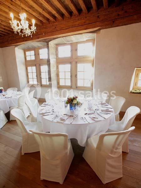 Salle lumineuse avec tables rondes dressées, nappes blanches, chaises houssées et lustre suspendu au plafond en bois.