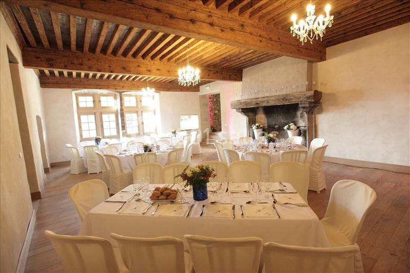 Salle de réception avec tables dressées, chaises blanches, cheminée en pierre et plafond en bois avec lustres.