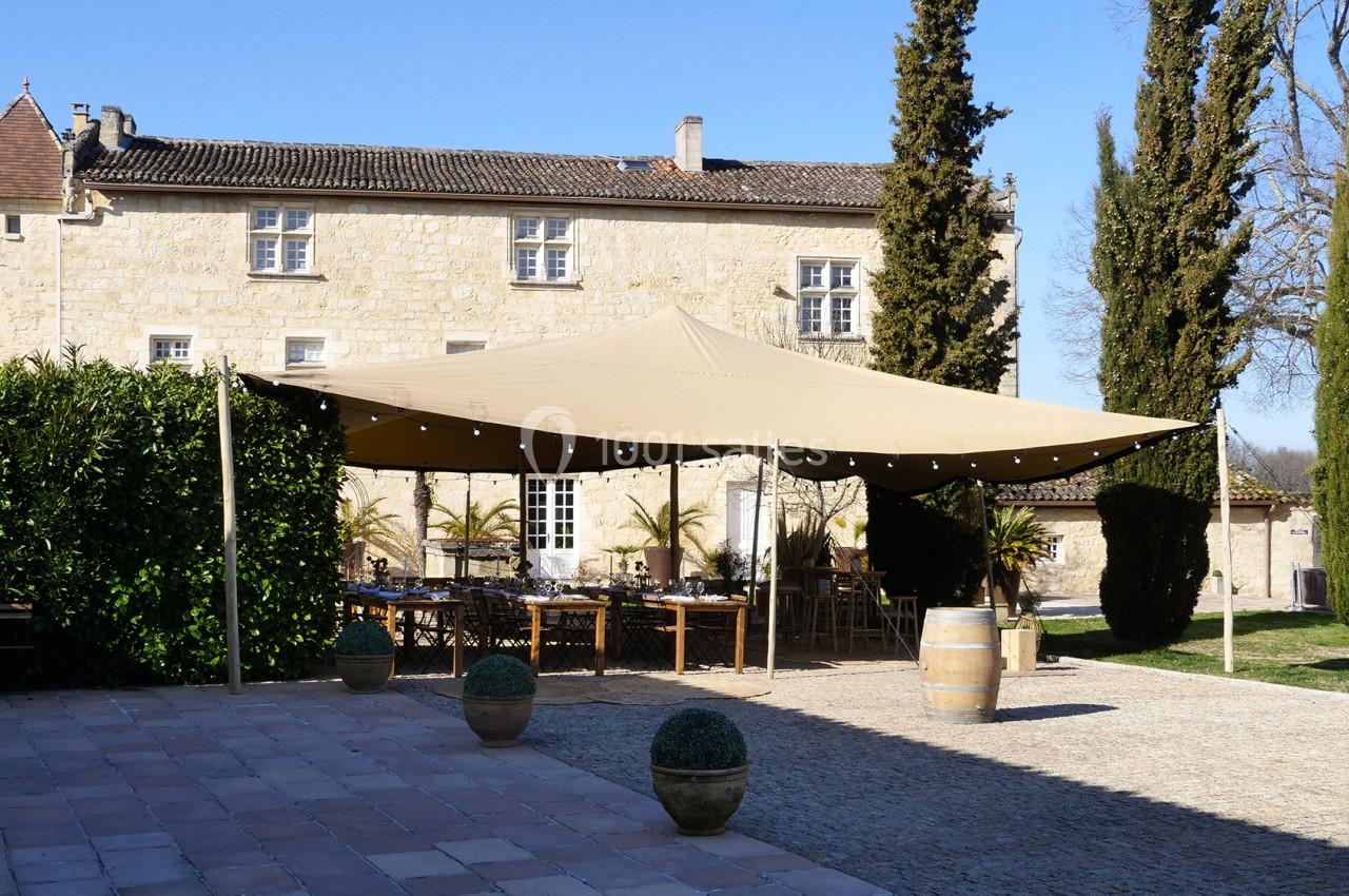 Grande tente beige installée dans une cour pavée devant un bâtiment en pierre, entourée de cyprès et de verdure.