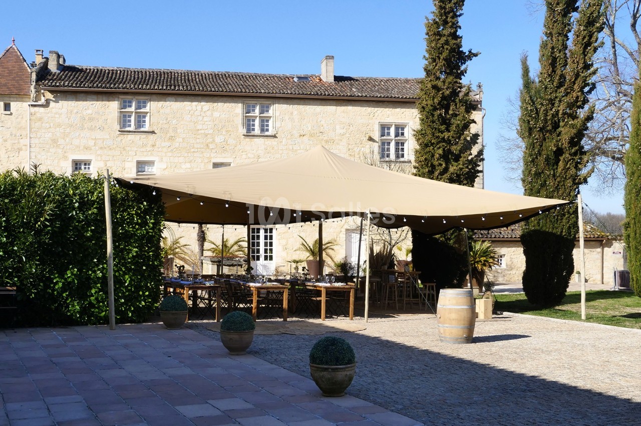 Grande tente beige installée dans une cour pavée devant un bâtiment en pierre, entourée de cyprès et de verdure.