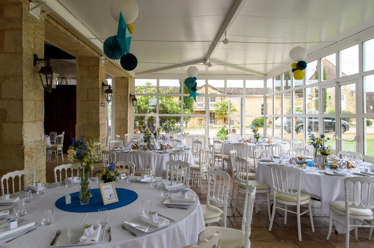 Salle de réception lumineuse avec tables rondes décorées de nappes blanches, fleurs et lanternes suspendues.