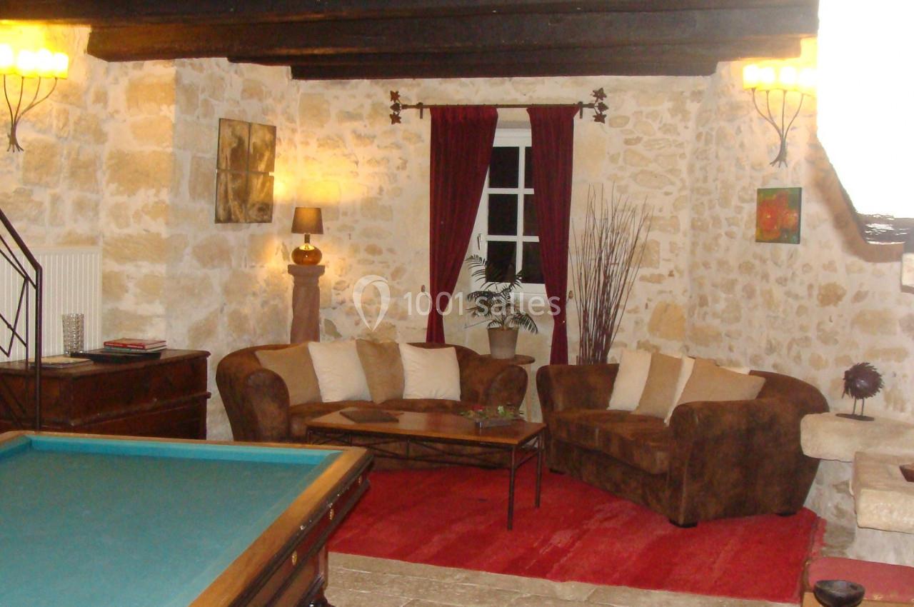 Salon rustique avec murs en pierre, canapé en velours marron, table basse, tapis rouge et table de billard au premier plan.