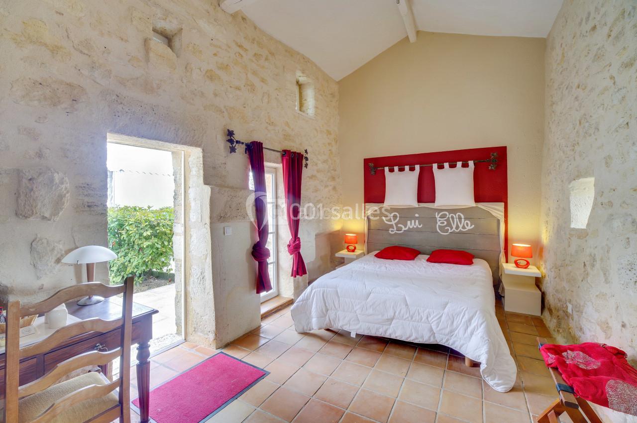 Chambre lumineuse avec murs en pierre, lit double, rideaux rouges et vue sur un jardin par une porte ouverte.