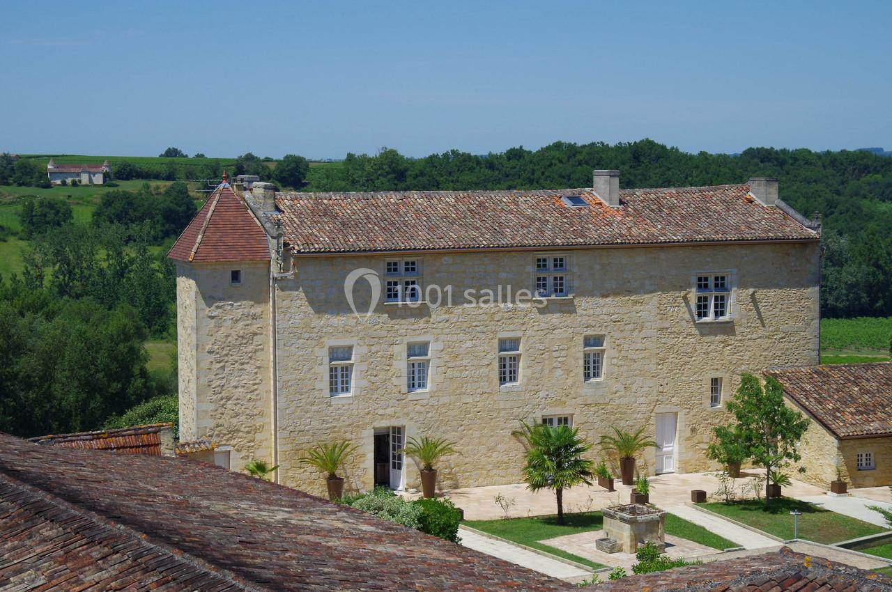 Bâtisse en pierre avec toit en tuiles, entourée de verdure et d'une cour avec des palmiers, sous un ciel bleu.