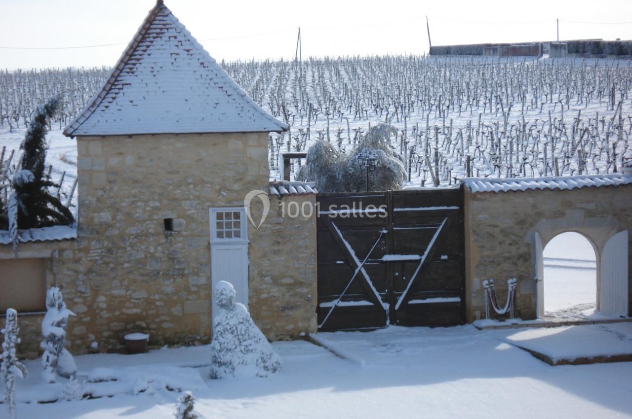 Cour enneigée avec un bâtiment en pierre, une porte en bois et des vignes recouvertes de neige en arrière-plan.