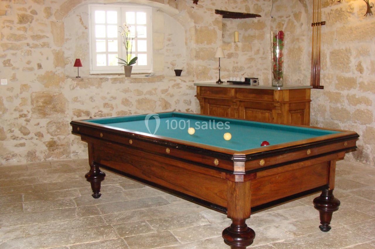 Table de billard en bois dans une pièce en pierre avec une fenêtre, un meuble et des queues de billard accrochées au mur.