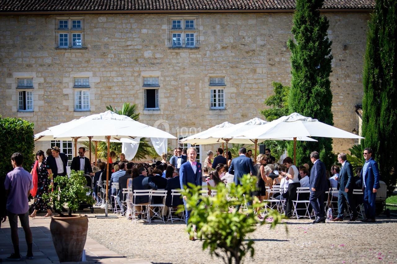 Groupe de personnes assises sous des parasols dans une cour pavée devant un bâtiment en pierre.