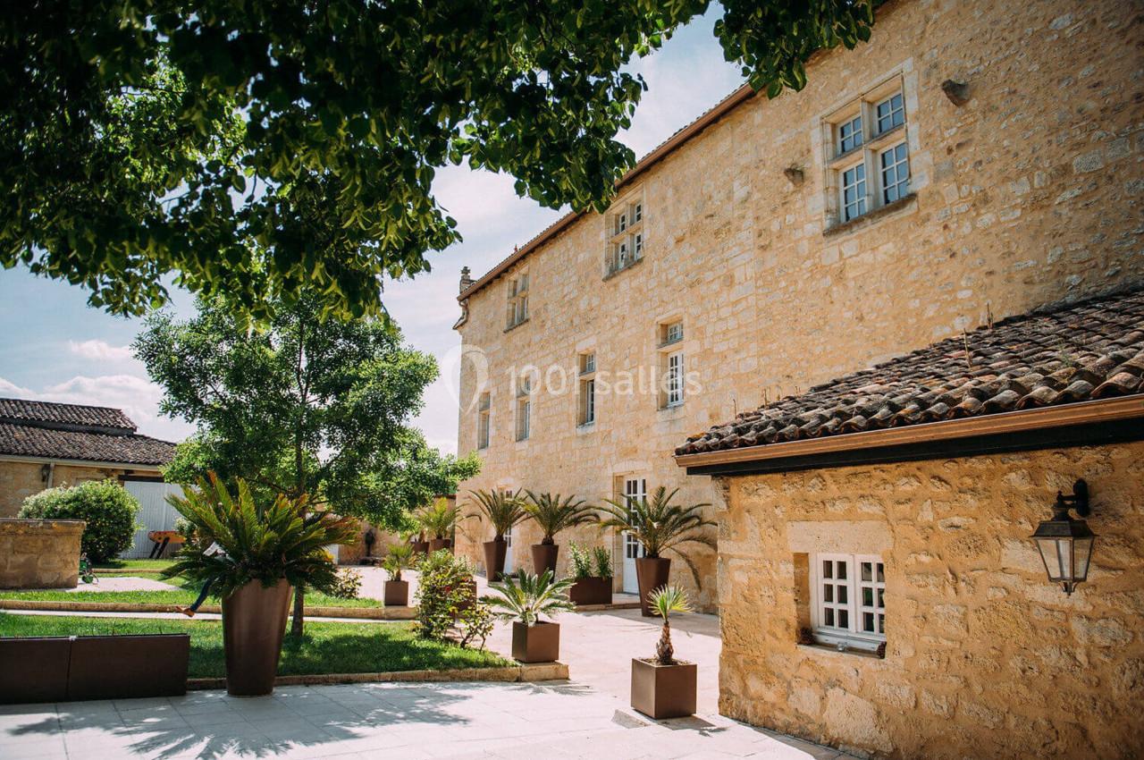 Façade en pierre d'un bâtiment ancien entouré d'arbres, de plantes en pots et d'une cour pavée.
