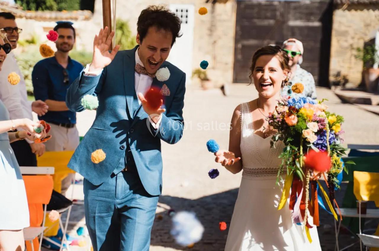 Un couple de mariés souriants traverse une allée extérieure sous des jets de pompons colorés.