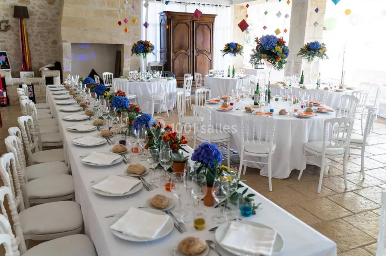 Salle de réception décorée pour un événement, avec tables dressées, nappes blanches et arrangements floraux colorés.
