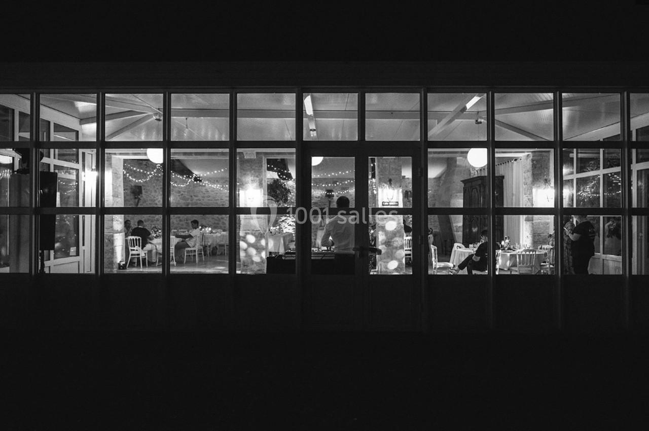 Vue nocturne d'une salle éclairée avec des baies vitrées, des tables décorées et quelques personnes à l'intérieur.