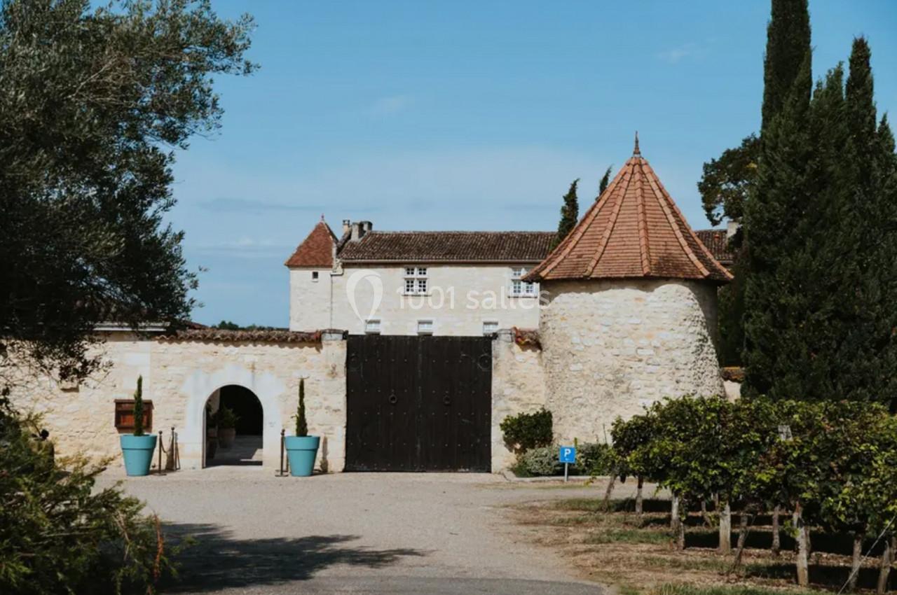 Bâtiment en pierre avec une tour à toit conique, portail en bois, arbres et vignes en premier plan.