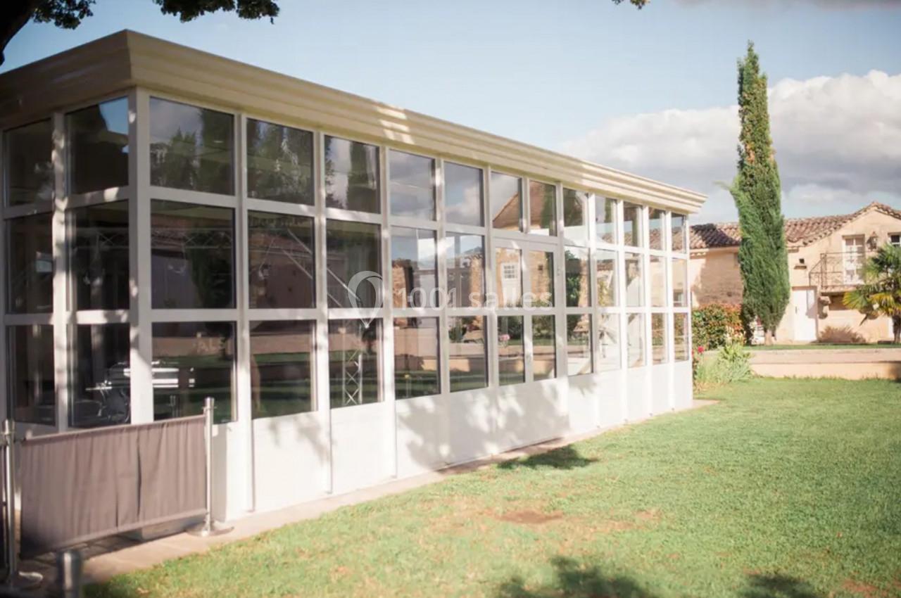 Structure vitrée moderne avec des murs en verre, entourée de pelouse et d'arbres, sous un ciel partiellement nuageux.