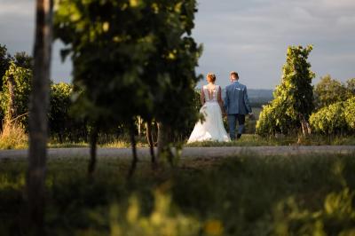 Une mariée en robe blanche marche sur une terrasse en bois, un enfant ajuste son voile sous un ciel dégagé.