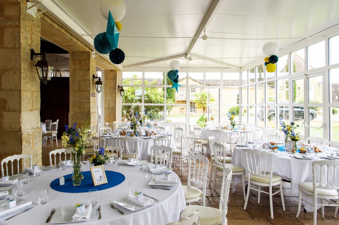 Salle lumineuse décorée pour un événement, avec des tables rondes dressées, des fleurs et des suspensions colorées.
