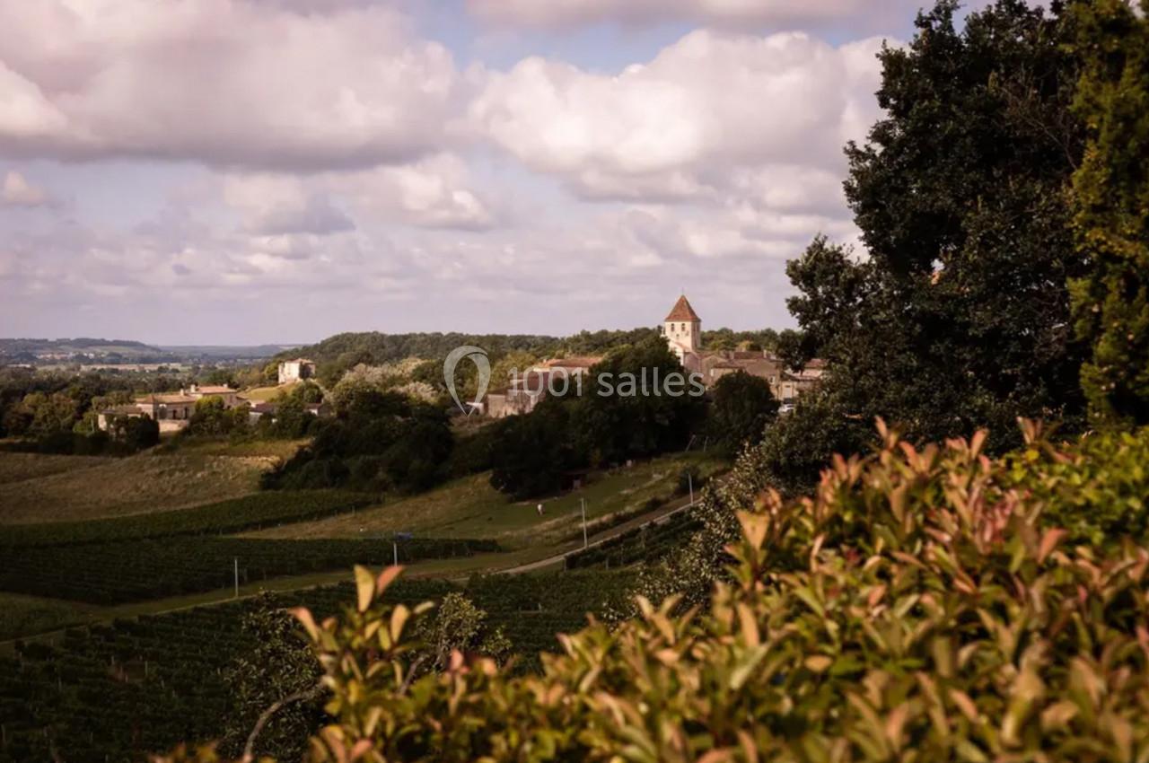 Village entouré de vignes et de verdure sous un ciel partiellement nuageux, avec une église visible au loin.